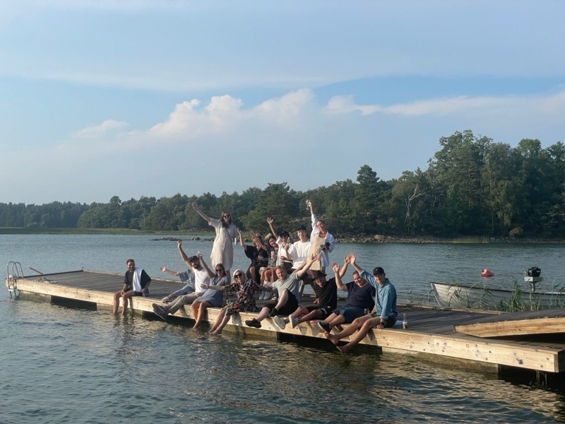 The Aimfor team on a jetty by the water on a summer evening, waving at the camera