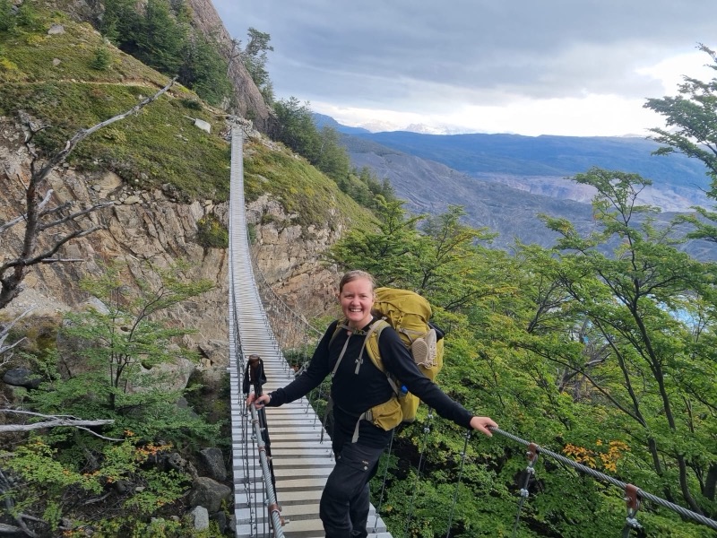 Annelie smiling at the start of a suspension bridge on a mountain hike, with a yellow backpack