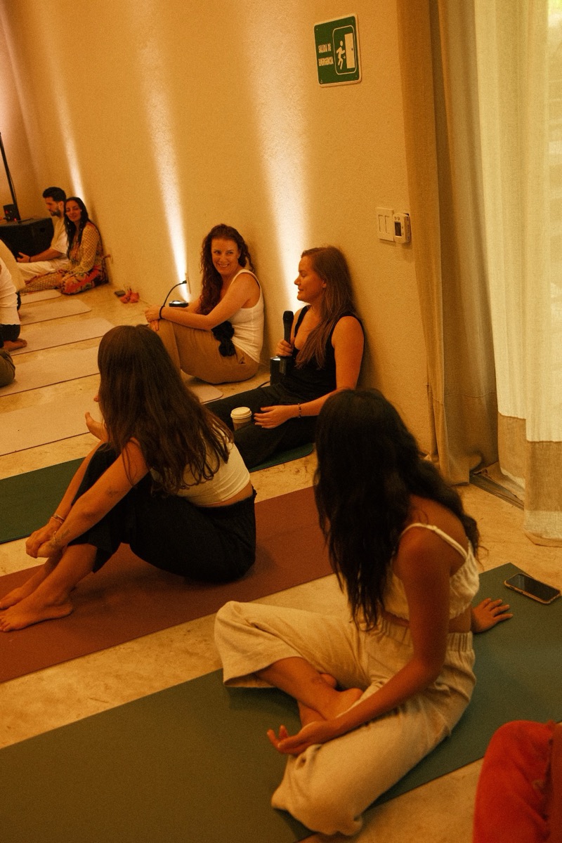 A group sitting on yoga mats in a softly lit room, Annelie holding a microphone and listening to a participant