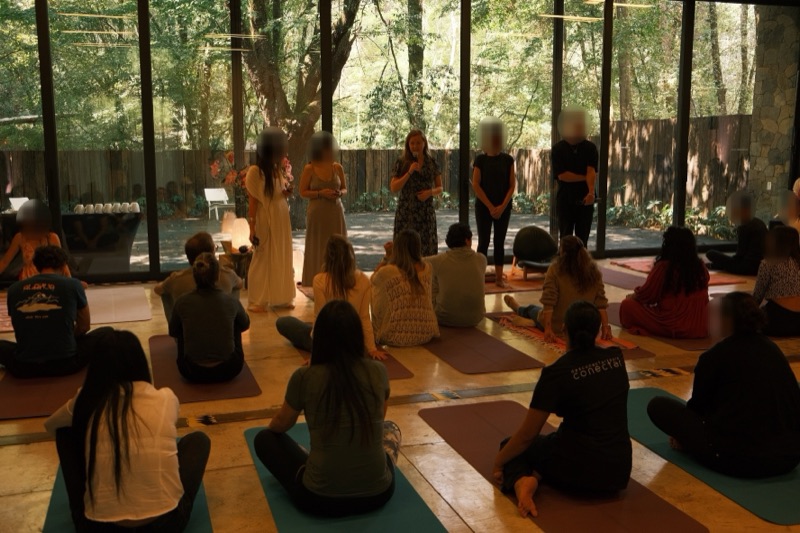 Annelie speaking into a microphone to a group seated on yoga mats, in a room with glass walls facing a forest