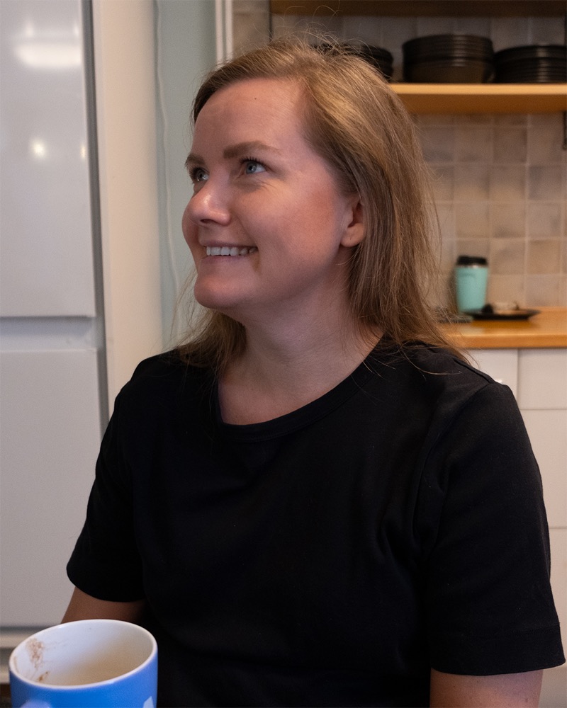 Annelie Näs in a kitchen with a mug in front of her, smiling and looking attentively to the side