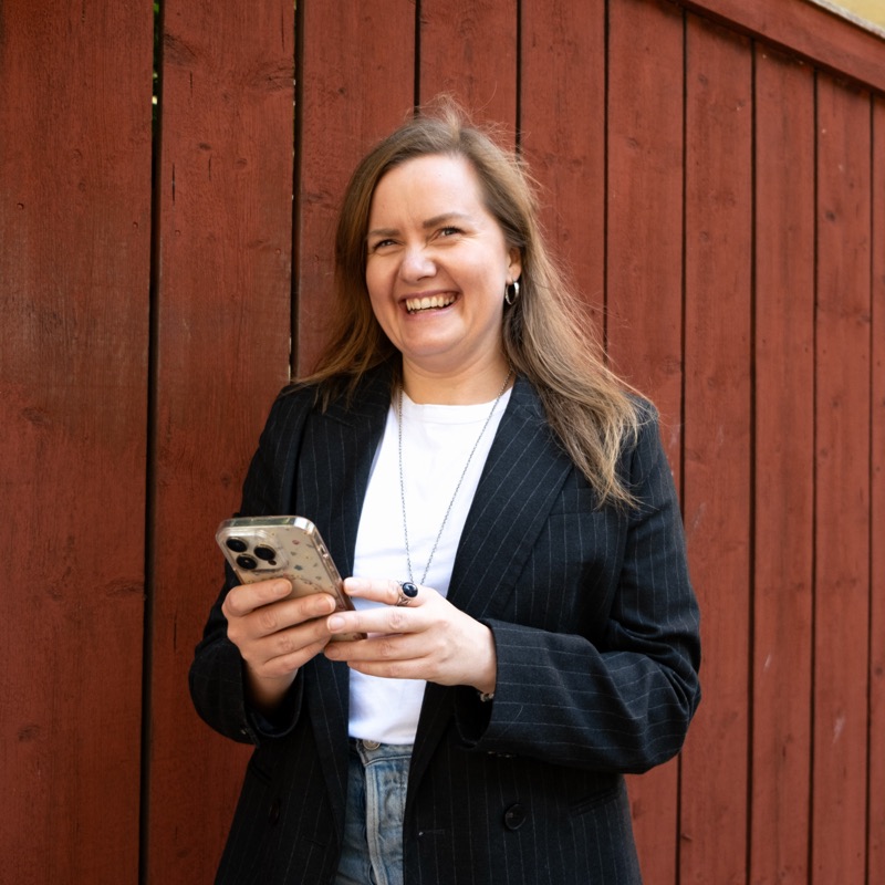 Annelie Näs skrattar framför en röd trävägg med telefonen i handen