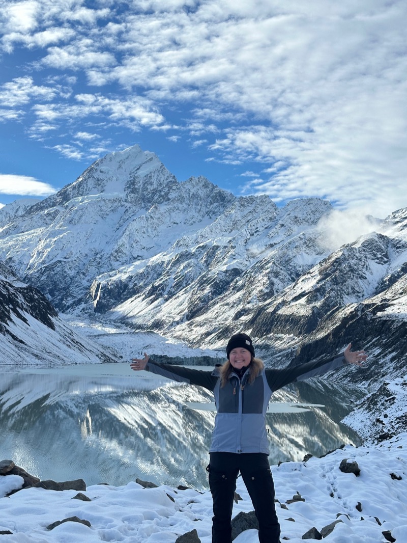 Annelie with her arms stretched wide in front of a snow-covered mountain and a glacier lake