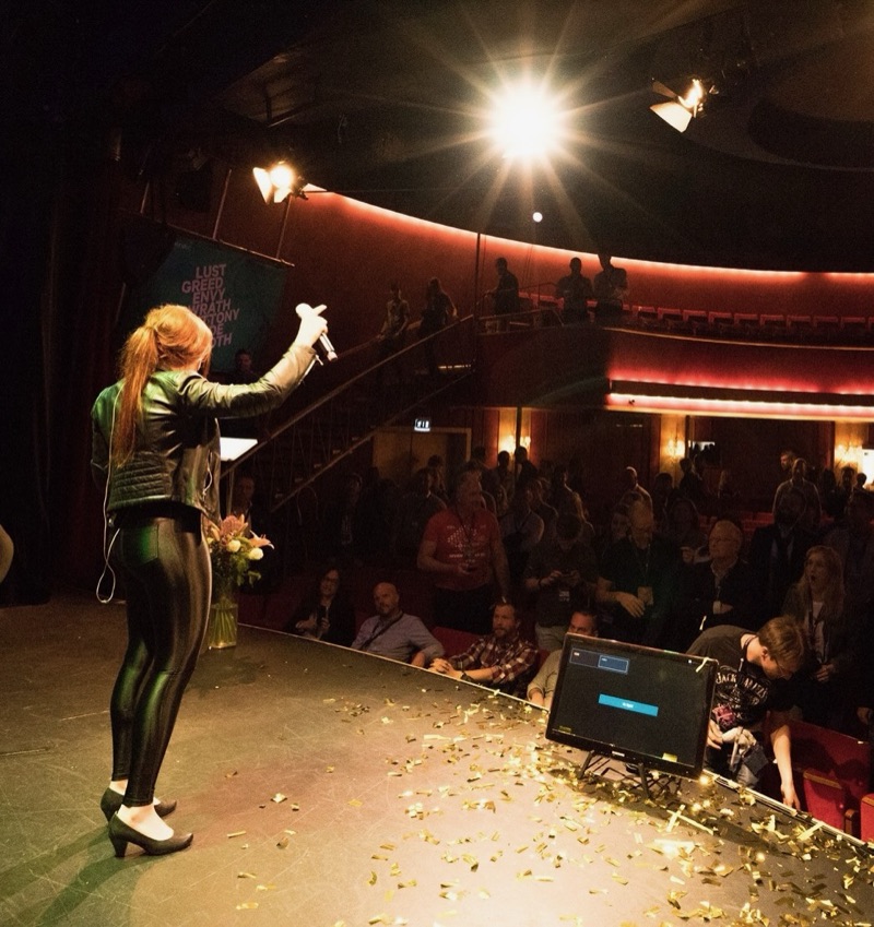 Annelie seen from behind on a theatre stage, microphone raised towards the audience, confetti on the floor