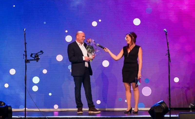 Annelie hosting a gala, holding out a microphone to a man with a bouquet of flowers