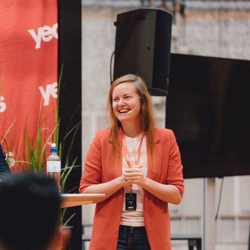 Annelie Näs laughing on stage at a conference, wearing a headset microphone and a red blazer