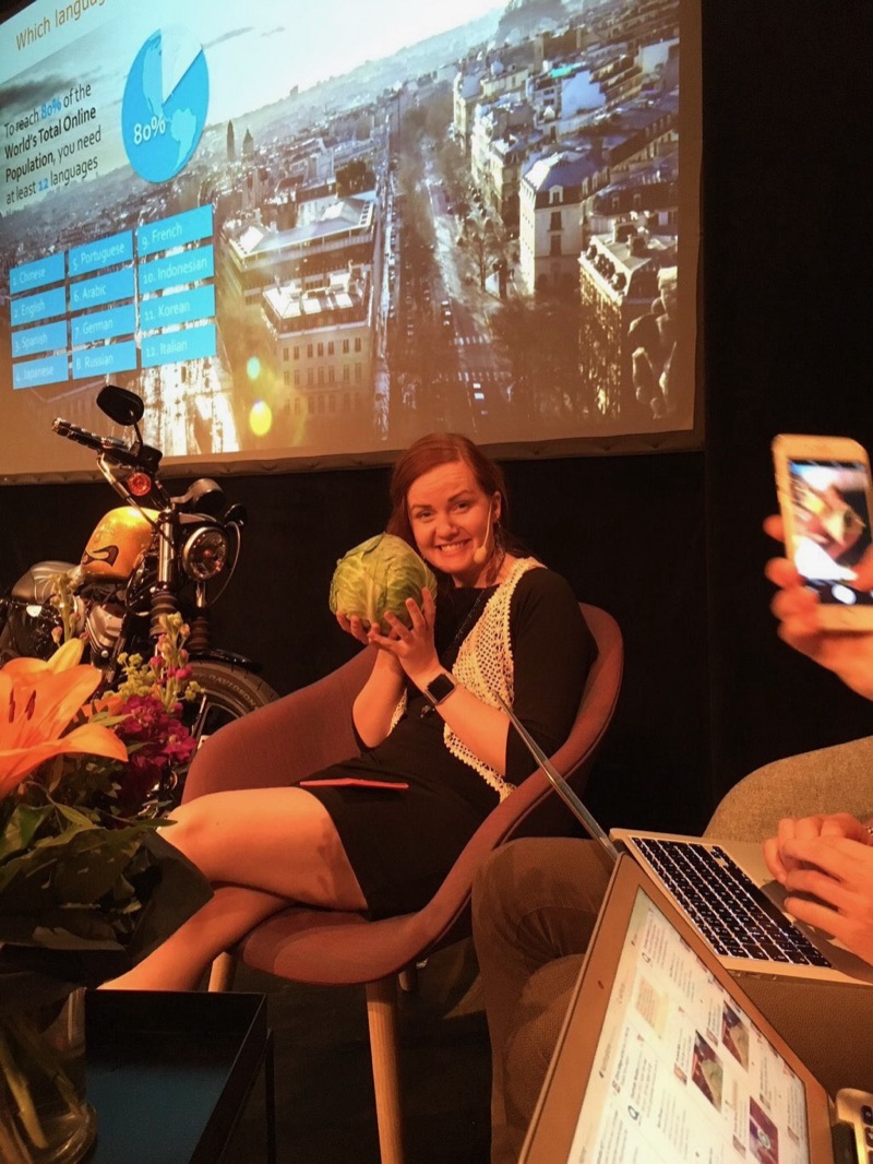 Annelie on stage in a moderator's chair with a headset, laughing and holding up a cabbage