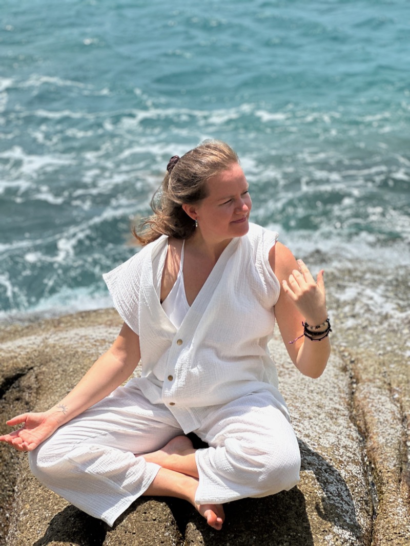 Annelie sitting cross-legged on a rock by the sea, dressed in white, looking out over the water