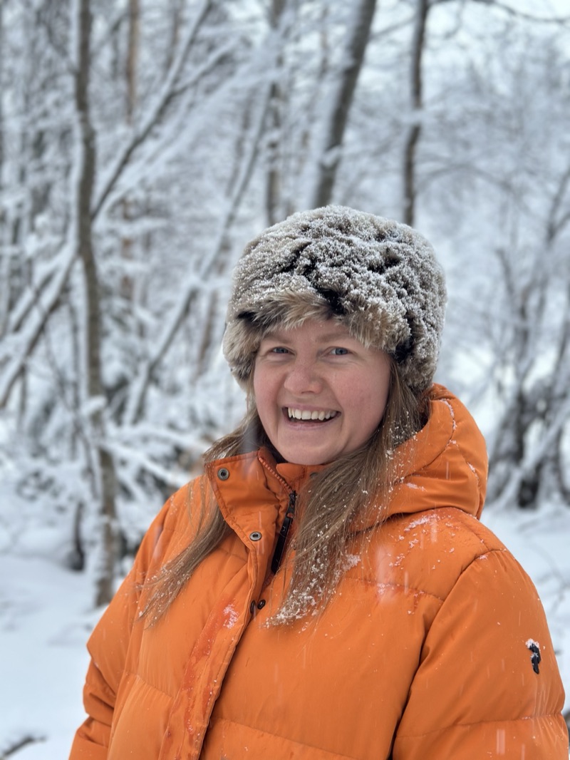 Annelie laughing in a snowy forest, wearing an orange down jacket and a fur hat dusted with snow