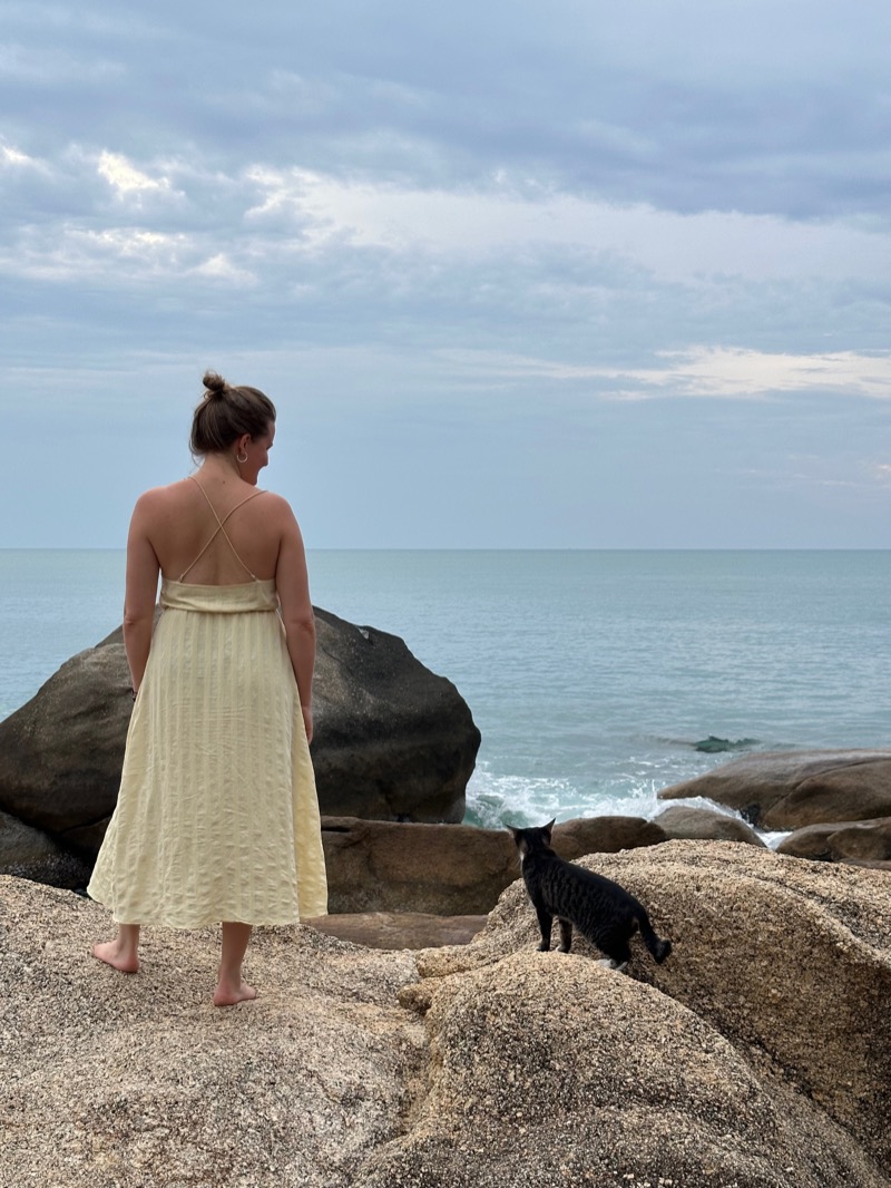 Annelie barefoot on a rock by the sea in a yellow dress, with a black cat beside her