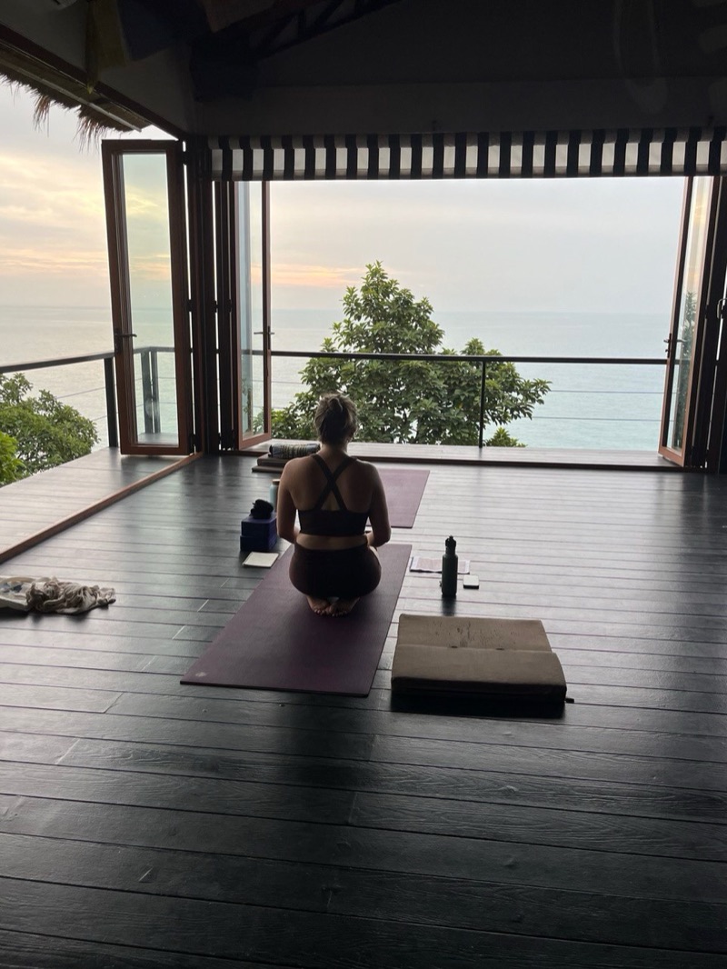 Annelie sedd bakifrån, på knä på en yogamatta i ett öppet rum mot havet i gryningen