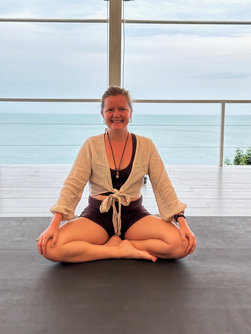 Annelie sitter med benen i kors på en yogamatta och ler, med havet bakom sig